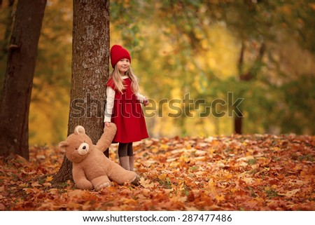 little smiling girl in red hat and red dress standing in the autumn park with teddy bear