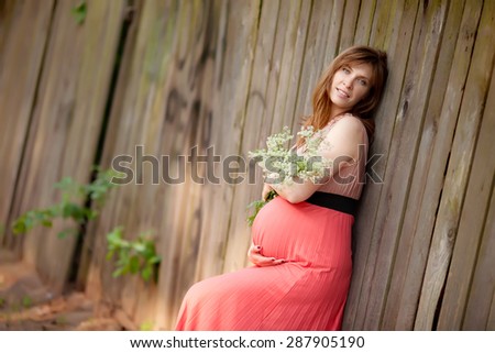 Beautiful smiling pregnant woman with long hair standing near the fence with white flowers in her hands at summer time