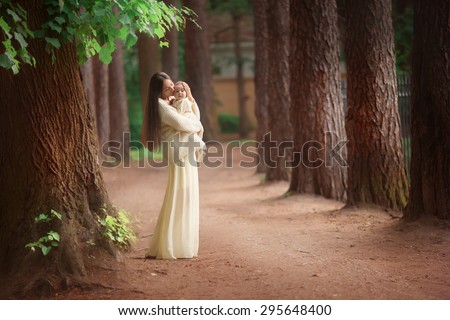 mother in long yellow dress is standing on the path in the park or forest near the trees and holding her baby girl daughter on her hands hugging and kissing her