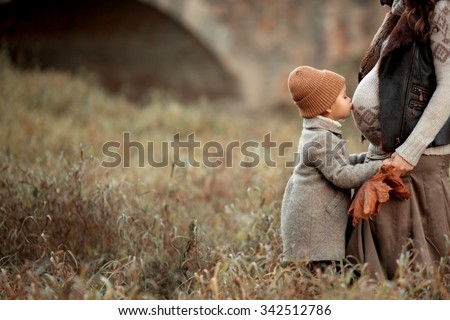 pretty little girl in brown hat and coat standing in the meadow in the park and kiss stomach of her pregnant mother at autumn