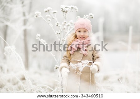 pretty little smiling girl with long blond hair in pink knitted hat and scarf and beige coat standing in the white snowy park or forest at winter time
