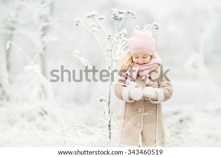 pretty little smiling girl with long blond hair in pink knitted hat and scarf and beige coat standing in the white snowy park or forest at winter time