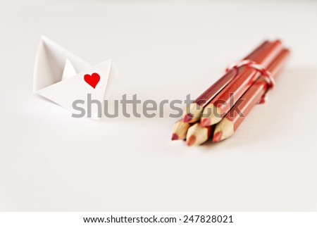 White paper boat with a red heart drawn on one side and a bunch of red color pencils