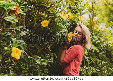 Blonde girl in pink t-shirt stay near small tree with yellow flowers and smell it