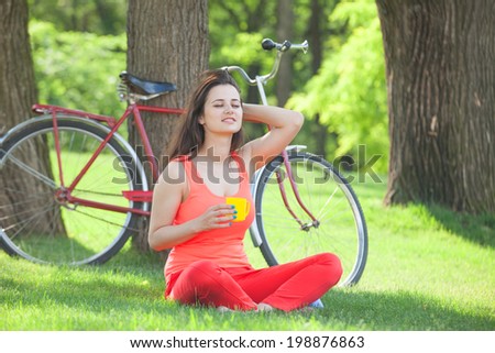 Brunette girl with cup of coffee in the park and with bicycle on background.