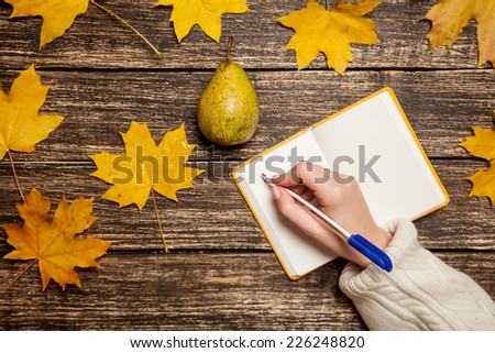 Female hand writing something in to notebook near pear on a table.
