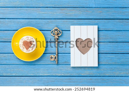 Cup of Cappuccino with heart shape symbol, key and photo frame on blue wooden background.