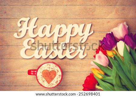 Cup of Cappuccino with heart shape symbol and words Happy Easter near flowers on wooden background