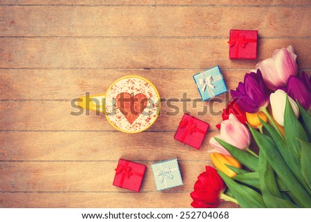 Cup of Cappuccino with heart shape symbol and gifts near flowers on wooden background