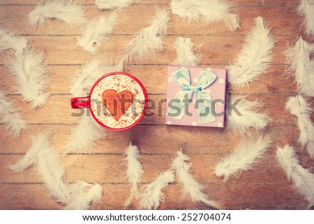 Cup of Cappuccino with heart shape symbol and feathers with gift on wooden background