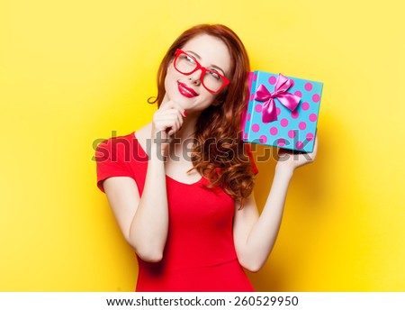 Redhead girl in red dress with glasses and gift box on yellow background
