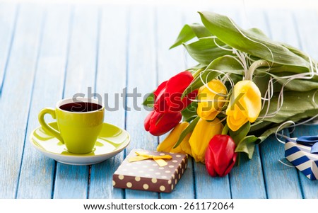 Cup of coffee and gift box with bouquet of tulips on blue table.