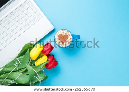 Cup of cappuccino with heart shape and computer, tulips on blue background.