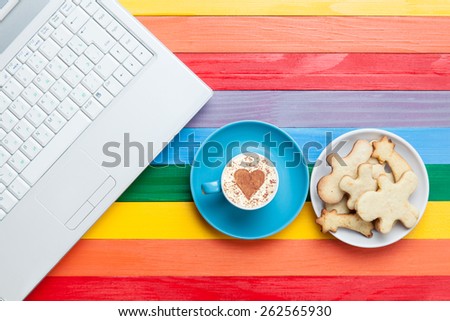Cup of cappuccino with heart shape and computer on multicolor wooden background.