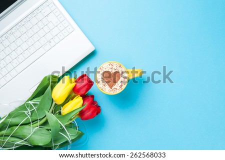 Cup of cappuccino with heart shape and computer, tulips on blue background.