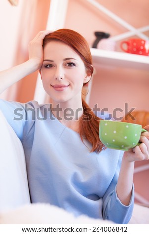 Redhead girl in blue blouse with green cup sitting on sofa at home