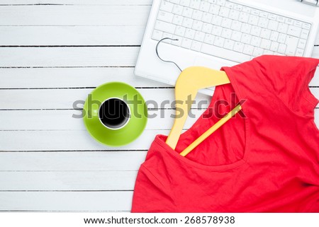 Cup of coffee and hanger with red dress near computer on white wooden table.