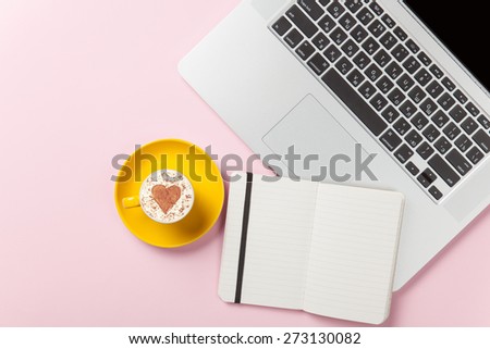 Cup of cappuccino with heart shape and laptop with note on pink background.