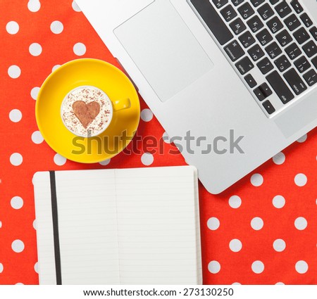 Cup of cappuccino with heart shape and laptop with note on red polka dot background.