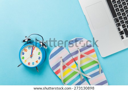 Alarm clock with flip flops near notebook on blue background
