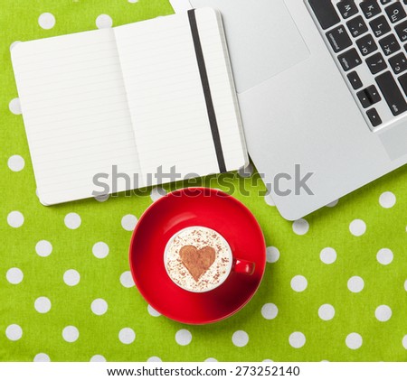 Cup of cappuccino with heart shape and laptop with note on green polka dot background.