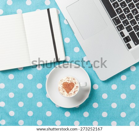 Cup of cappuccino with heart shape and laptop with note on blue polka dot background.