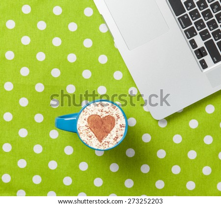 Cup of cappuccino with heart shape and laptop on green polka dot background.