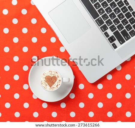 Cup of cappuccino with heart shape and laptop on red polka dot background.