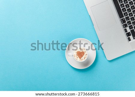 Cup of cappuccino with heart shape and laptop on blue background.