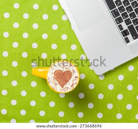 Cup of cappuccino with heart shape and laptop on green polka dot background.