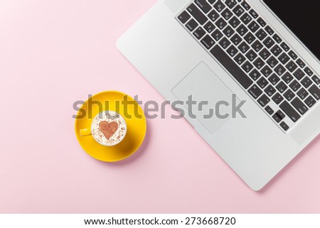Cup of cappuccino with heart shape and laptop on pink background.
