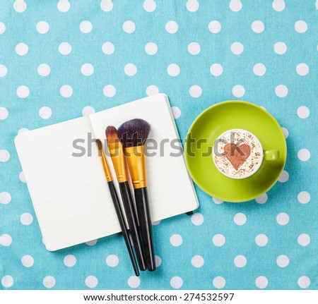 Cup of cappuccino with heart shape and notebook with makeup brushes on blue polka dot background.