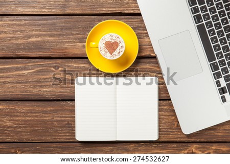 Cup of cappuccino with heart shape and notebook near laptop on wooden table.