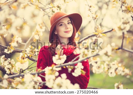Portrait of a beautiful redhead women in red sweater and hat with cup in blossom apple tree garden in spring time on sunset.