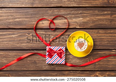 Cup of cappuccino with heart shape and red ribbon  near gift box on wooden background.