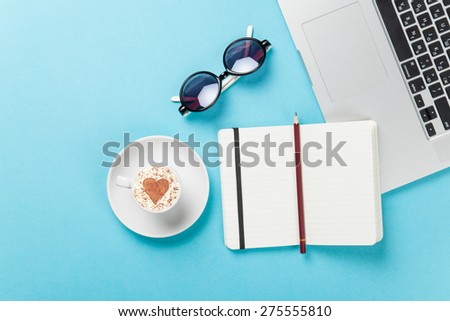 Cup of cappuccino with heart shape and laptop near glasses on blue background.