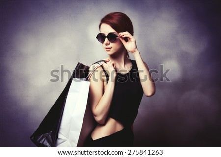 Style redhead girl with sunglasses and black dress with shopping bags on grey background