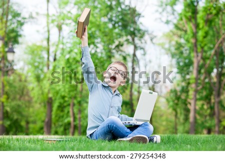 young surprised boy with a books and laptop computer on green grass in the park