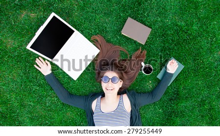 portrait of a beautiful brunette student girl with laptop computer and books on the green grass in the park in spring time.