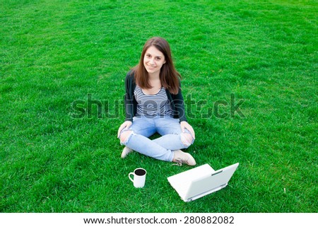 portrait of a beautiful brunette student girl with laptop computer on the green grass in the park in spring time.