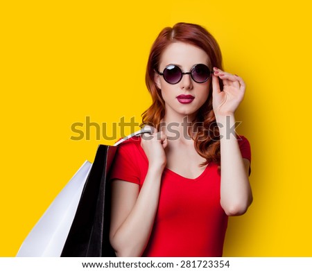 Redhead girl in red dress and sunglasses with shopping bags on yellow background