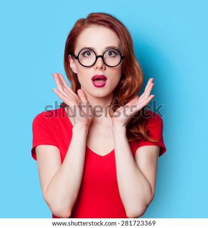 Surprised redhead girl in red dress with glasses on blue background
