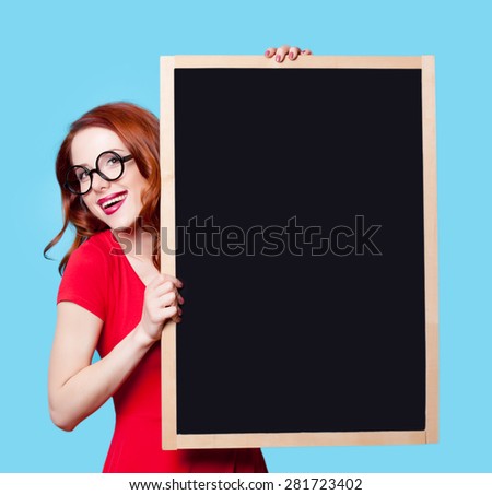 Young smiling redhead student in red dress with blackboard on blue background.