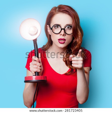 Surprised redhead girl in red dress with lamp on blue background