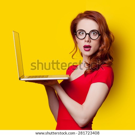 Surprised redhead girl with laptop computer in red dress on yellow background.