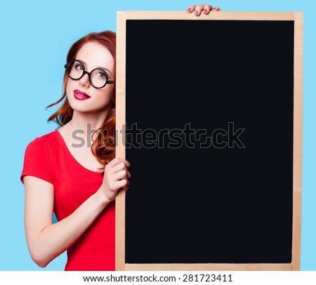 Young surprised redhead student in red dress with blackboard on blue background.