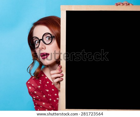 Young surprised redhead teacher in red polka dot dress with blackboard on blue background.