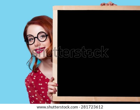 Young smiling redhead teacher in red polka dot dress with blackboard on blue background.