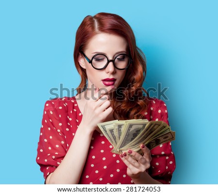 Surprised redhead girl in red polka dot dress with money on blue background.