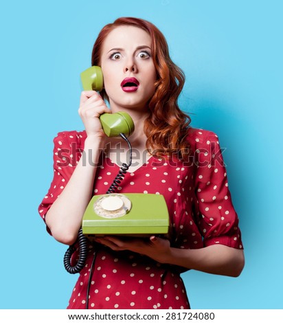 Surprised redhead girl in red polka dot dress with green dial phone on blue background.